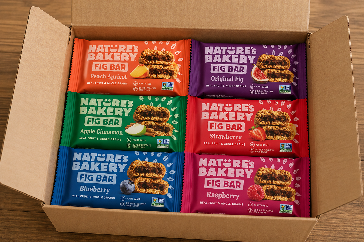 Nature’s Bakery Fig Bars variety pack arranged inside an open plain craft shipping box featuring Peach Apricot, Original Fig, Apple Cinnamon, Strawberry, Blueberry, and Raspberry snack bars on a wooden table.