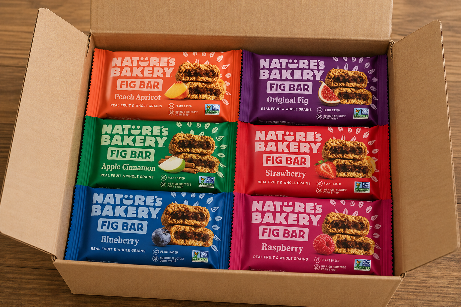 Nature’s Bakery Fig Bars variety pack arranged inside an open plain craft shipping box featuring Peach Apricot, Original Fig, Apple Cinnamon, Strawberry, Blueberry, and Raspberry snack bars on a wooden table.
