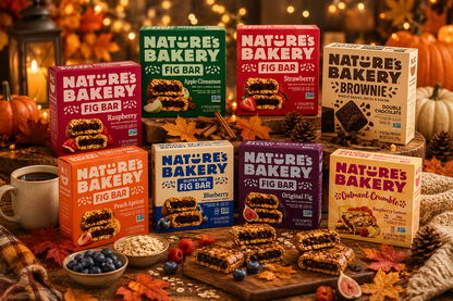 Nature’s Bakery fall snack display featuring fig bars, oatmeal crumble bars, and brownie bars with pumpkins, autumn leaves, coffee, and cozy seasonal décor on a rustic wooden table.